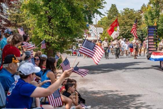 Spectators line a tree-lined neighborhood street waving American flags as a patriotic parade passes by. Scouts carrying flags lead the procession while families watch from lawn chairs and the curb under a sunny summer sky.