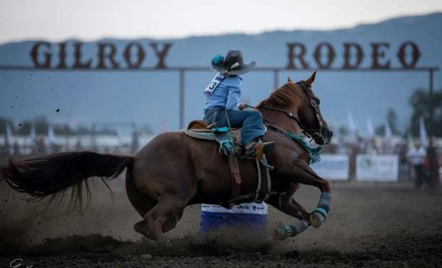 a cowboy riding a brown horse with the Gilroy Rodeo sign in the background