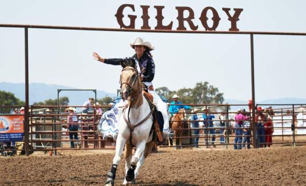 A young lady in a white cowboy horse waving rides an shite and brown horse with the Gilroy rodeo sign behind her