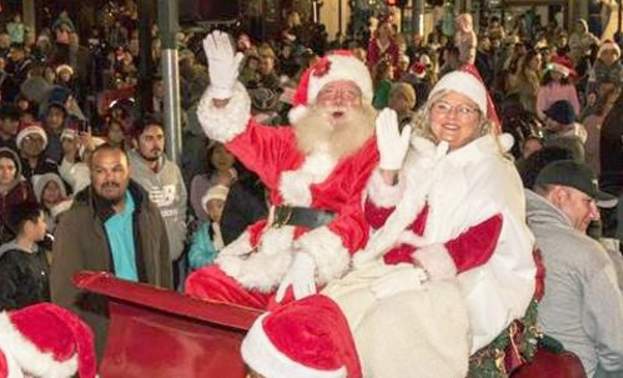 a man and women dressed in red and white as Santa and Mrs. Claus riding in a red sleigh in a parade with people all around