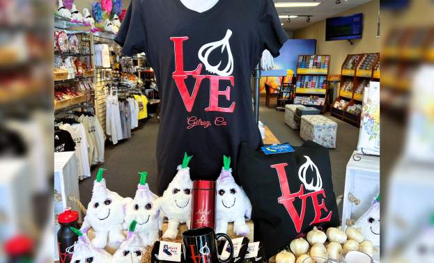 Table display of gift shop items with garlic on them - plush garlic toys, Gilroy Garlic socks, Gilroy logo beer mugs, and a "LOVE Garlic" Gilroy California T-shirt.