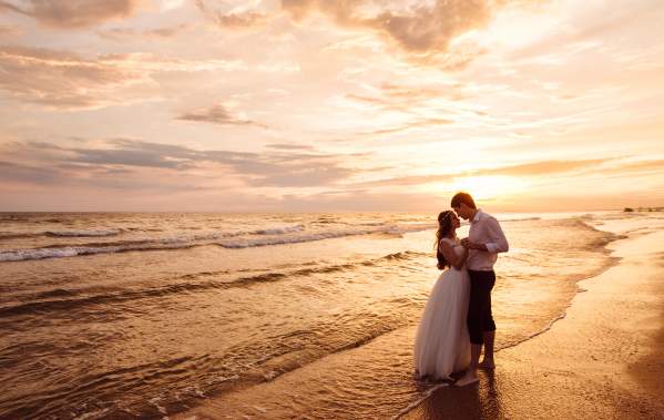A couple embracing on the beach at evening in their wedding attire.