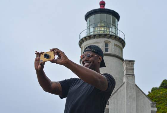 A person stands in front of the Heceta Lighthouse and holds up a camera.