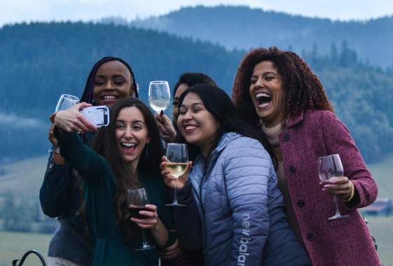 A group of people holding wine glasses lean in together with a cell phone for a group selfie.