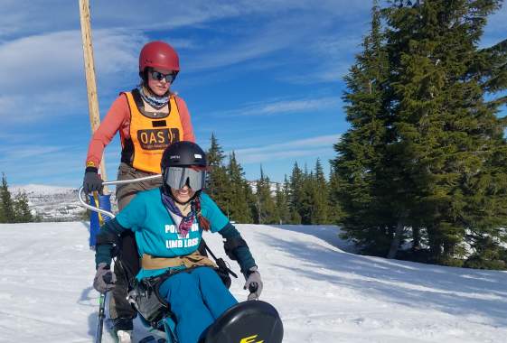 A skier uses adaptive ski equipment with a companion to ski the slopes. Snow, trees and blue sky in the backdrop.