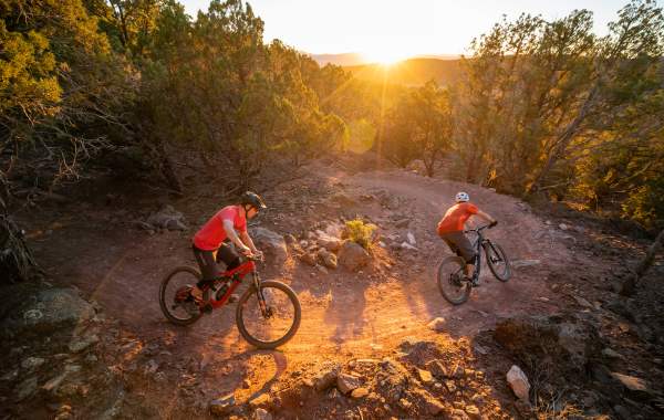 Two cyclists rounding a turn on the Lichen It mountain bike trail in Cedar City Utah at sunset with a sunburst over the distant hills.