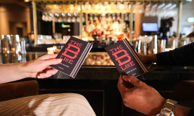 Two people's hands are are in the foreground, holding paper B-Line Guides. In the background is a bar with many bottles of bourbon.