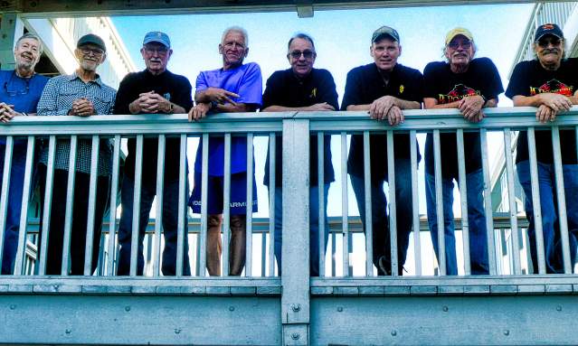 A group of eight men lean against a balcony and smile down at the camera