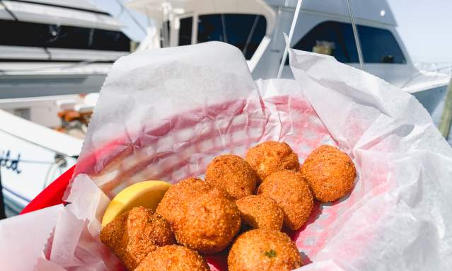 A basket of hushpuppies is being held up in front of a large white yacht on the water.