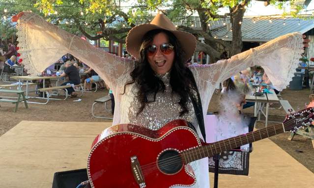 A woman in a white outfit with a red guitar holds her arms out in celebration