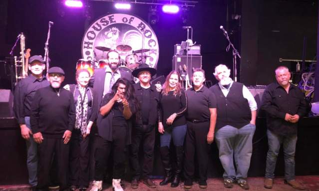 A group of 10 band members stand in front of a stage set up with band equipment.