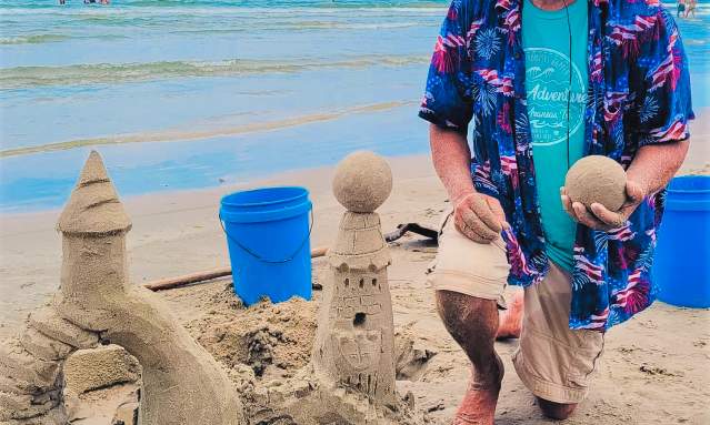 A smiling man kneels next to a sandcastle on the beach. He holds a ball of sand in one hand.