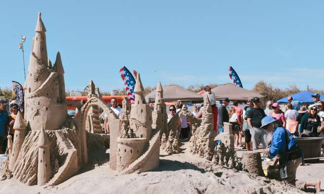 In the foreground, the back of a sandcastle is visible. In the background, the sculptor kneels over his project while a crowd watches and strolls by.