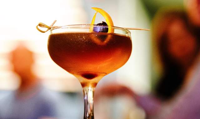 A close-up of a cocktail in a stemmed glass, garnished with an orange twist and cherry, with blurred figures in the background.