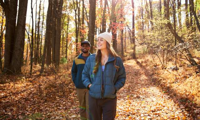 A couple walks on a trail in a forest with fallen brown and orange leaves wearing warm jackets and hats on a fall day.