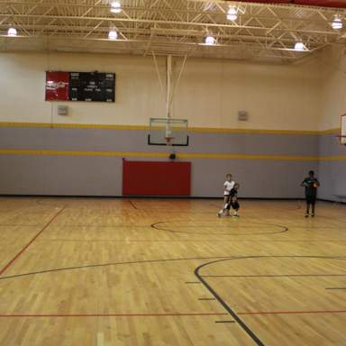 Indoor basketball gym with hardwood floors, multiple hoops, and overhead lighting, where three children stand near center court during practice.