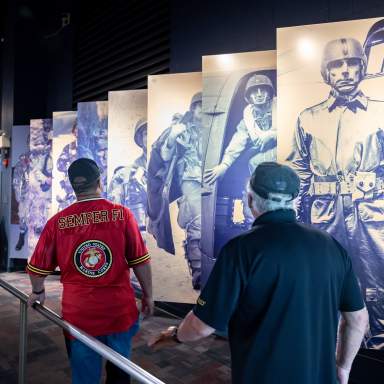 Two visitors walk through a military history exhibit lined with large blue-toned panels of paratroopers and nearby display cases at Fayetteville's ASOM.