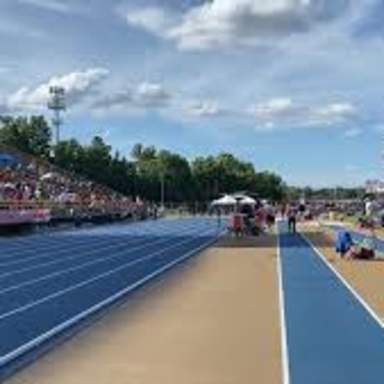 A wide view of the Cape Fear High School track during a meet, featuring bright blue lanes, athletes warming up, and spectators filling the stadium bleachers under a partly cloudy sky.