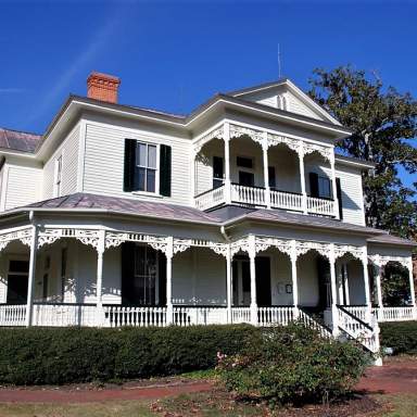 The 1897 Poe House, a two-story white Victorian home in Fayetteville, NC, featuring an ornate wraparound porch, decorative trim, and an upper balcony under a bright blue sky.
