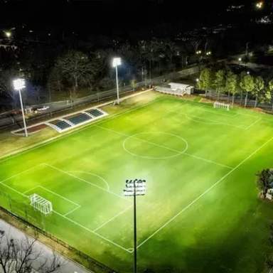 Aerial nighttime view of the Duggins Soccer Stadium fully lit with bright field lights, green turf, and seating areas surrounding the pitch.