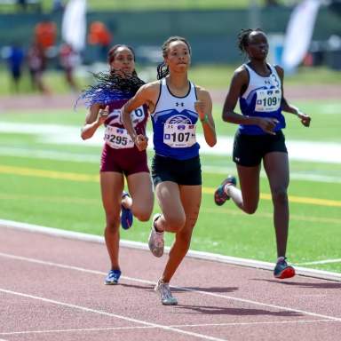 Three female track athletes run on the FSU outdoor track during a race, with the lead runner in a blue and white uniform and two competitors close behind on a sunny day.