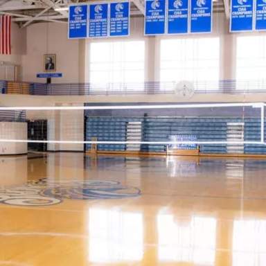 Indoor volleyball court at Fayetteville State University with polished hardwood floors, blue padded posts, and bleachers in the background beneath championship banners.