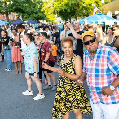Diverse group of people dancing and smiling at a Fourth Friday event in downtown Fayetteville, NC, with vendor tents and summer sunlight in the background.