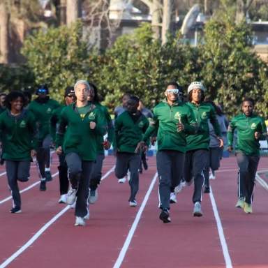 A large group of Methodist University track athletes jogs together on an outdoor track, wearing matching green warm-up jackets, with trees and training equipment visible in the background.
