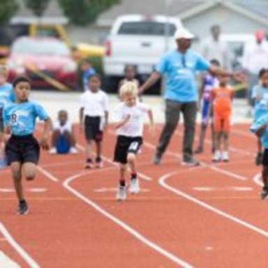 Young children sprint down a red running track during a youth race, with coaches and other participants watching from the sidelines.