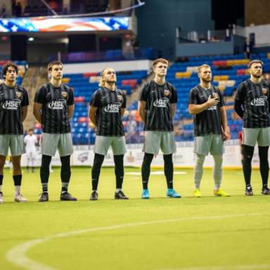 Indoor soccer players from the Fayetteville Fury line up on the field before a match, standing shoulder to shoulder as they face the audience, with one player holding his hand over his heart.