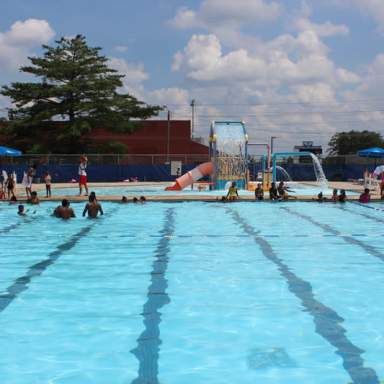 Outdoor swimming pool at Smith Recreation Center with lane markings, families swimming, and a splash-play structure and slide in the background on a sunny day.