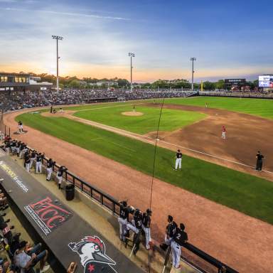 Wide view of a baseball stadium at sunset with a game in progress and fans filling the stands