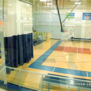 Indoor gymnasium with a hardwood basketball court, blue and red floor markings, retractable divider curtain, and bleacher seating along the sidelines.