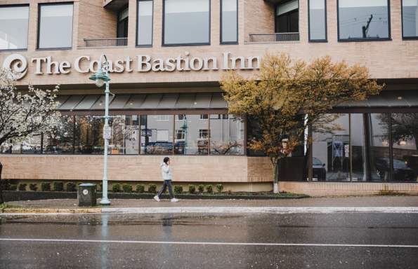 A person walks past The Coast Bastion Inn on a rainy day, with wet pavement and trees lining the street.