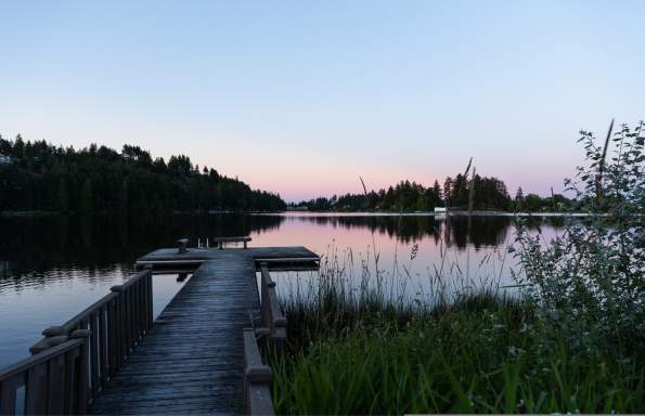 A quiet wooden dock leads out over a still lake at dusk, with soft pink and blue tones in the sky.
