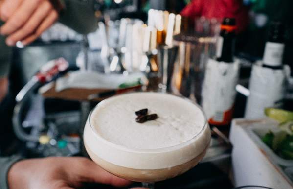 A hand holding a creamy espresso martini garnished with coffee beans at a lively bar, with blurred colorful lights and bartending tools in the background.