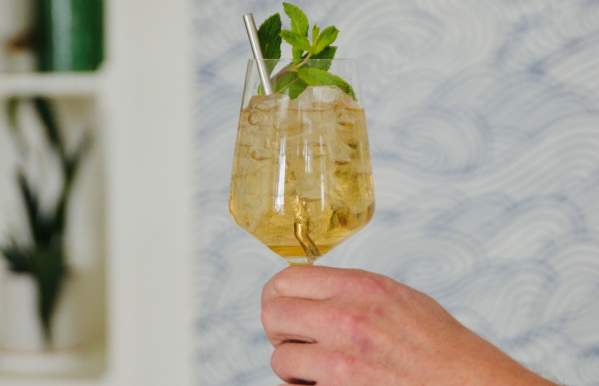 A hand holding a wine glass filled with a golden cocktail, garnished with fresh mint and a metal straw, against a stylish blue and white patterned background.