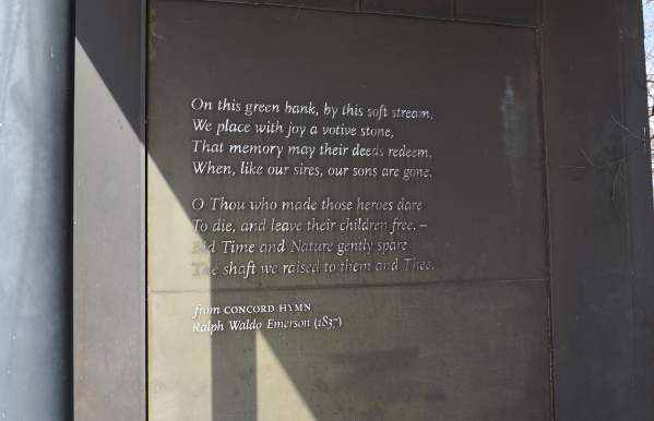 A close-up image of a bronze plaque mounted on a dark metal wall at the Grounds of Remembrance in Dublin, Ohio. The plaque features an excerpt from Concord Hymn by Ralph Waldo Emerson (1837), engraved in elegant serif text. Sunlight casts a triangular shadow across the lower portion of the plaque, emphasizing the engraved words. The poem honors fallen heroes and speaks of memory, sacrifice, and freedom.