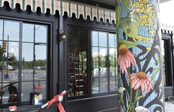 A colorful mosaic-covered pillar featuring large flowers like sunflowers and coneflowers stands outside a building with black-framed windows and a striped awning. A vintage red scooter is parked on the brick sidewalk in front of the pillar, creating a vibrant and artistic streetscape.