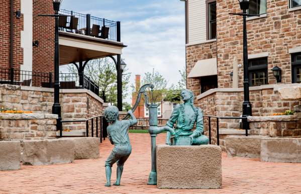 Bronze sculpture of a child pumping water for a seated Native American woman in a brick plaza surrounded by historic buildings.