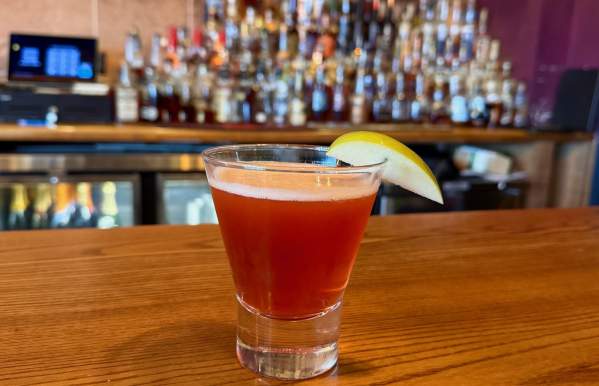 A vibrant red cocktail served in a short glass, garnished with a fresh apple slice, sits on a wooden bar counter. The blurred background showcases an array of liquor bottles on shelves, creating a warm and inviting bar atmosphere.