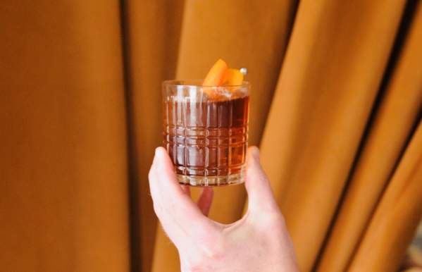 A hand holding a classic whiskey cocktail in a textured glass, garnished with an orange peel, against a backdrop of warm, golden-brown curtains.