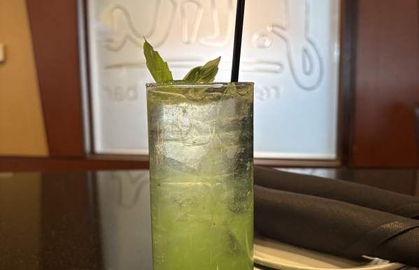A tall glass of vibrant green cocktail with ice, garnished with fresh basil leaves and a black straw, sits on a dark countertop. In the background, a frosted glass window with the word 'Live' is visible, along with a neatly folded black napkin on a white plate.
