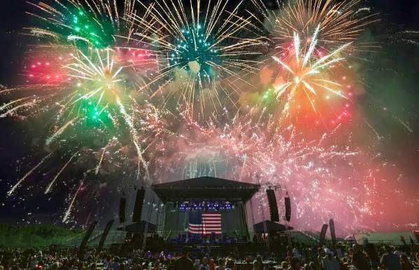 A colorful Four of July fireworks display in the sky above a stage that has the American flag hanging as the backdrop