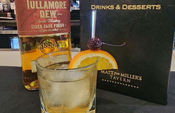 A glass of whiskey on the rocks garnished with an orange slice and cherry, set on a black napkin. Behind it, a bottle of Tullamore D.E.W. Irish Whiskey and a 'Drinks & Desserts' menu from Matt the Miller’s Tavern are displayed on the bar