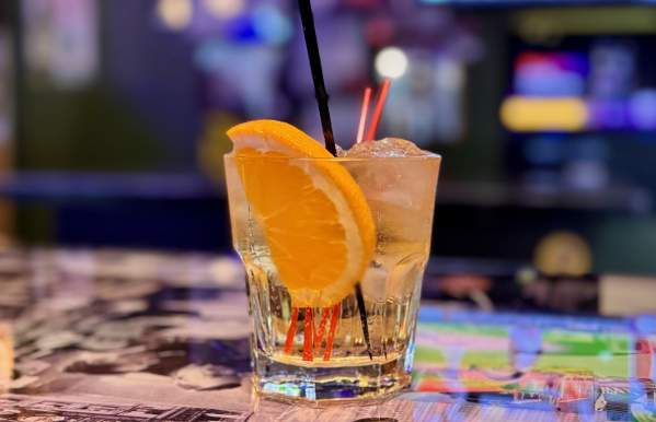 A classic cocktail served in a short glass over ice, garnished with a vibrant orange slice and black and red straws, sits on a bar counter covered in black-and-white photos. The blurred background features colorful bar lights and screens.