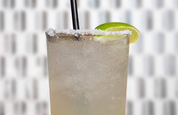 A salted-rim margarita in a tall glass filled with ice, garnished with a fresh lime wedge and a black straw, against a blurred geometric background.