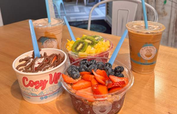 A wooden table with a display of Playa Bowls menu items, including an acai bowl topped with strawberries and blueberries, a dragon fruit bowl topped with kiwi and pineapple, a Nutella oatmeal bowl, and two beverages – an iced coffee and a smoothie.