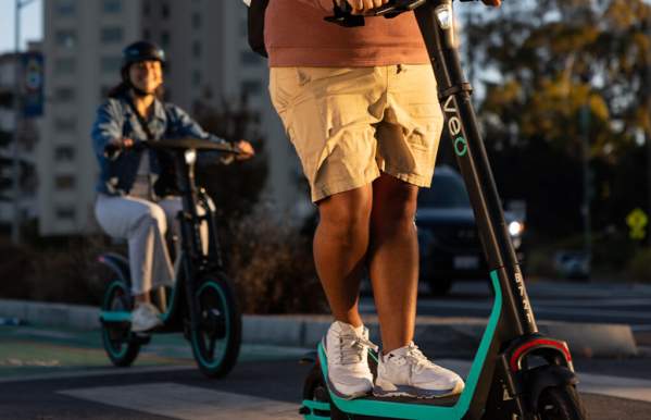Two people ride Veo electric scooters and bikes on a city street, enjoying a sunny day with traffic and buildings in the background.