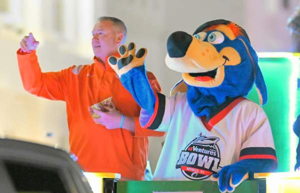 A mascot dressed as a blue dog in a football jersey and a football coach in an orange shirt wave to the crowd from a Mardi Gras float